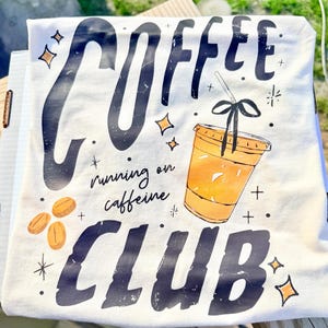 May include: A white t-shirt with a coffee-themed graphic. The design includes the word "COFFEE" in large, distressed black letters, a coffee cup, coffee beans, and the phrase "running on caffeine CLUB".