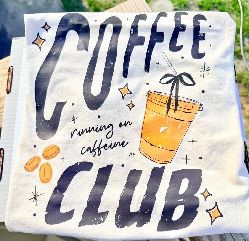 May include: A white t-shirt with a coffee-themed graphic. The design includes the word "COFFEE" in large, distressed black letters, a coffee cup, coffee beans, and the phrase "running on caffeine CLUB".