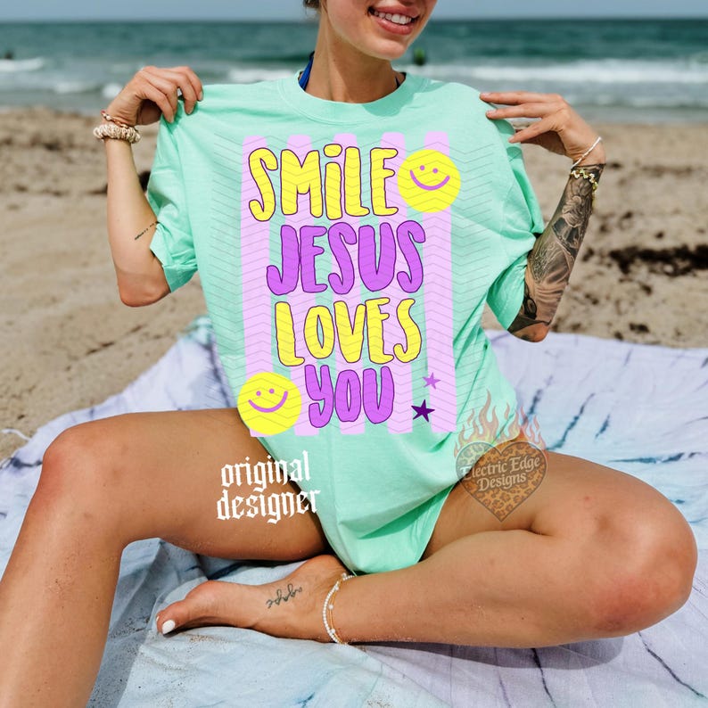 May include: A light green t-shirt with the words "SMILE JESUS LOVES YOU" in yellow and purple, along with smiley face graphics and a star. The shirt is worn on a beach.