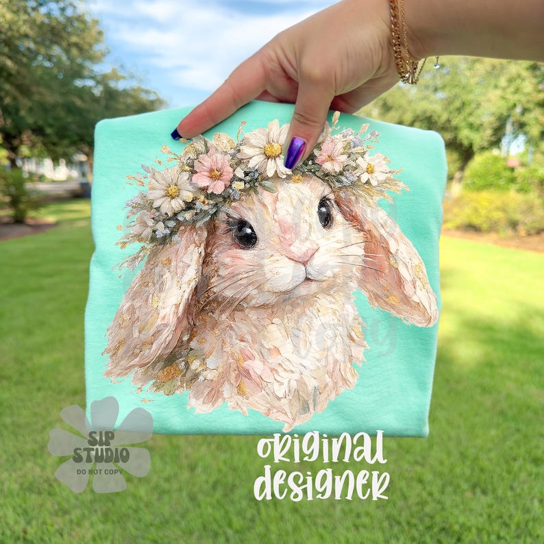 May include: A mint green t-shirt featuring a detailed illustration of a bunny wearing a floral crown. The bunny has soft, textured fur and large, expressive eyes. The text "original designer" is printed in white script on the shirt.