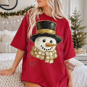 May include: A red short-sleeved t-shirt featuring a graphic of a snowman wearing a black top hat with a gold band and a matching gold and white checkered scarf. The snowman has a textured white finish, black eyes, a dotted mouth, and a gold nose.
