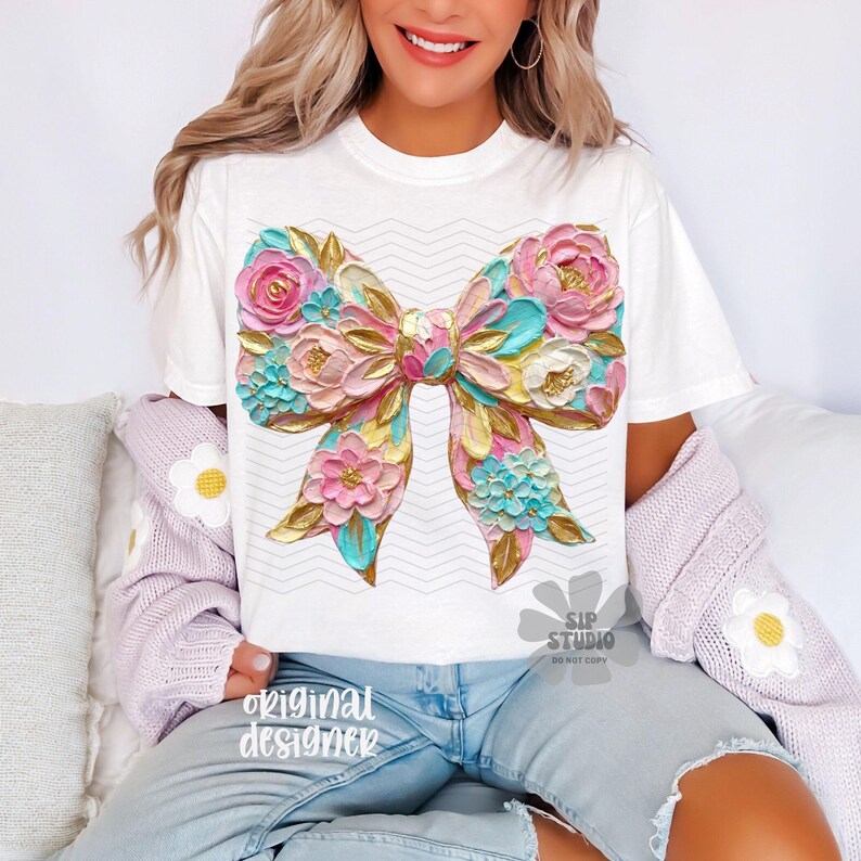 May include: White t-shirt featuring a large, colorful bow design with floral accents. The bow is rendered in shades of pink, blue, gold, and yellow, with a textured appearance. The shirt also has the text "original designer" at the bottom.