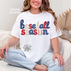 May include: White t-shirt with the words "baseball season" in blue, red, and cream-colored embroidered letters. The letter "n" is designed as a baseball. The shirt is worn by a person.