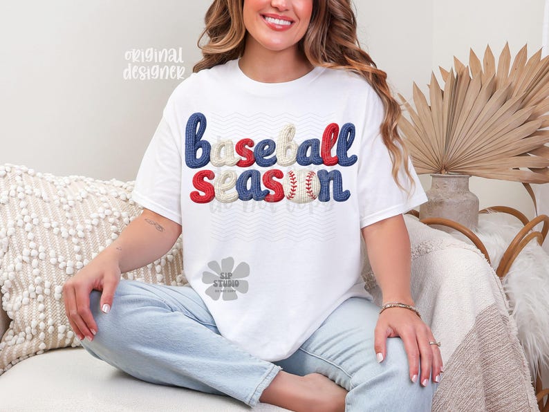 May include: White t-shirt with the words "baseball season" in blue, red, and cream-colored embroidered letters. The letter "n" is designed as a baseball. The shirt is worn by a person.