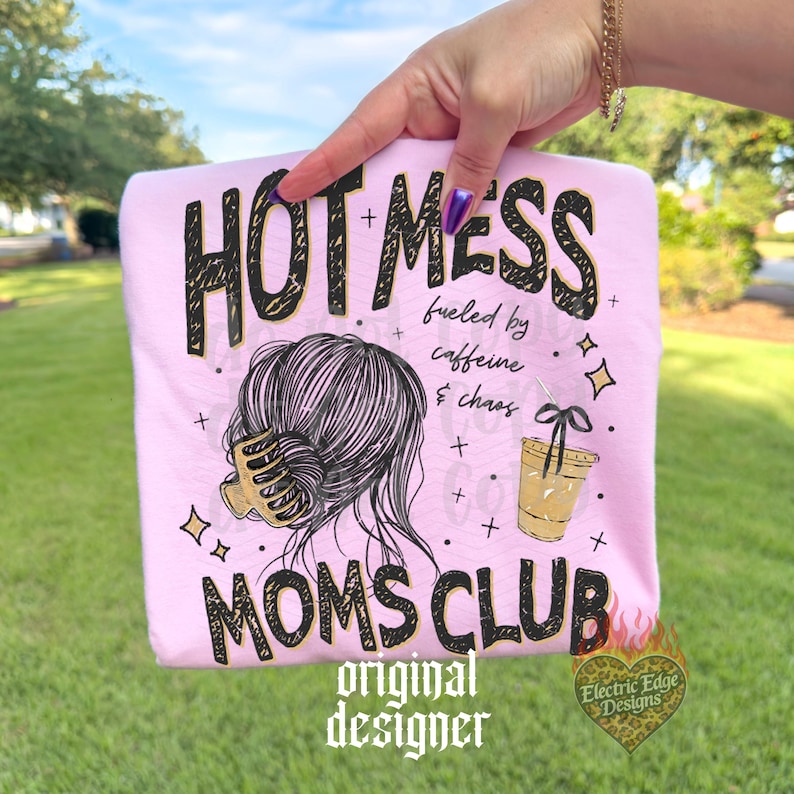 May include: A light pink t-shirt with the words "HOT MESS" in black and gold lettering. The shirt features a graphic of a woman's hair in a bun, a coffee cup, and the words "MOMS CLUB". The shirt is held up by a hand.