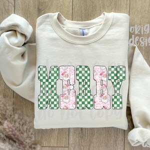 May include: Cream-colored sweatshirt with the word "MERRY" in large, block letters. The letters have a green and white checkered pattern with pink Santa Claus faces. The sleeves are rolled up.