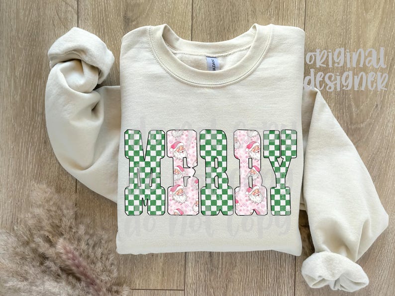 May include: Cream-colored sweatshirt with the word "MERRY" in large, block letters. The letters have a green and white checkered pattern with pink Santa Claus faces. The sleeves are rolled up.