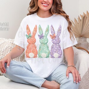 May include: White t-shirt featuring three pastel-colored Easter bunny designs. The bunnies are in shades of peach, mint green, and lavender, each with a bow and floral details. The text "original designer" is visible on the left side of the shirt.