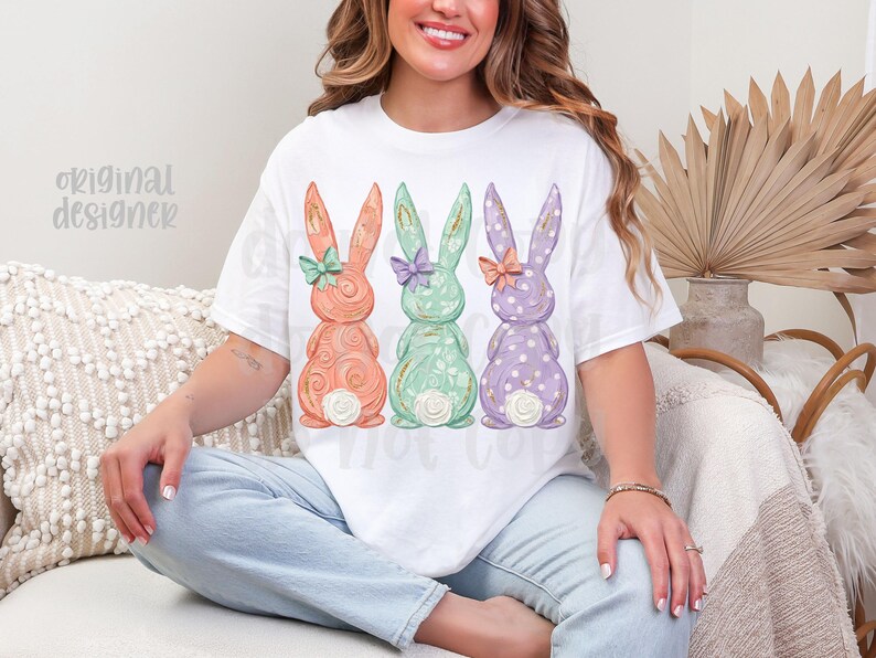 May include: White t-shirt featuring three pastel-colored Easter bunny designs. The bunnies are in shades of peach, mint green, and lavender, each with a bow and floral details. The text "original designer" is visible on the left side of the shirt.