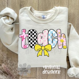 May include: Cream-colored sweatshirt with the word "TEACH" in a colorful, patchwork design. The letters feature stars, checkered patterns, lightning bolts, and smiley faces. A yellow bow is centered below the word.
