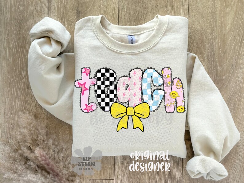 May include: Cream-colored sweatshirt with the word "TEACH" in a colorful, patchwork design. The letters feature stars, checkered patterns, lightning bolts, and smiley faces. A yellow bow is centered below the word.