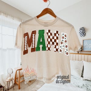 May include: A cream-colored t-shirt featuring the word MAMA in large block letters. The M is brown with white laces, the A is green glitter, the M is a brown and white checkerboard pattern, and the A is white with footballs and stars.