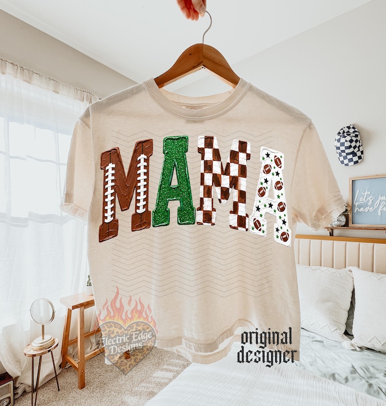 May include: A cream-colored t-shirt featuring the word MAMA in large block letters. The M is brown with white laces, the A is green glitter, the M is a brown and white checkerboard pattern, and the A is white with footballs and stars.