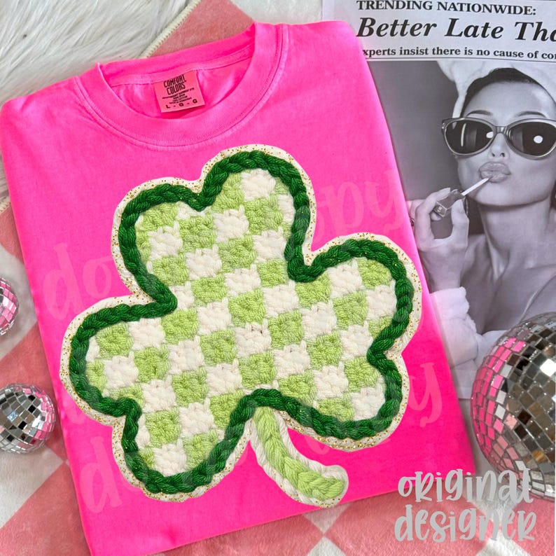 May include: A bright pink sweatshirt featuring a large, crocheted shamrock design. The shamrock has a green and white checkered pattern with a green border. The shirt has the Comfort Colors brand label.
