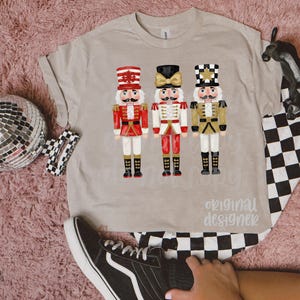 Peut inclure: T-shirt beige avec trois illustrations de casse-noisettes color&eacute;s. Les casse-noisettes portent des uniformes rouges, blancs et dor&eacute;s avec des bottes noires. Le texte "original designer" est imprim&eacute; sur le t-shirt.