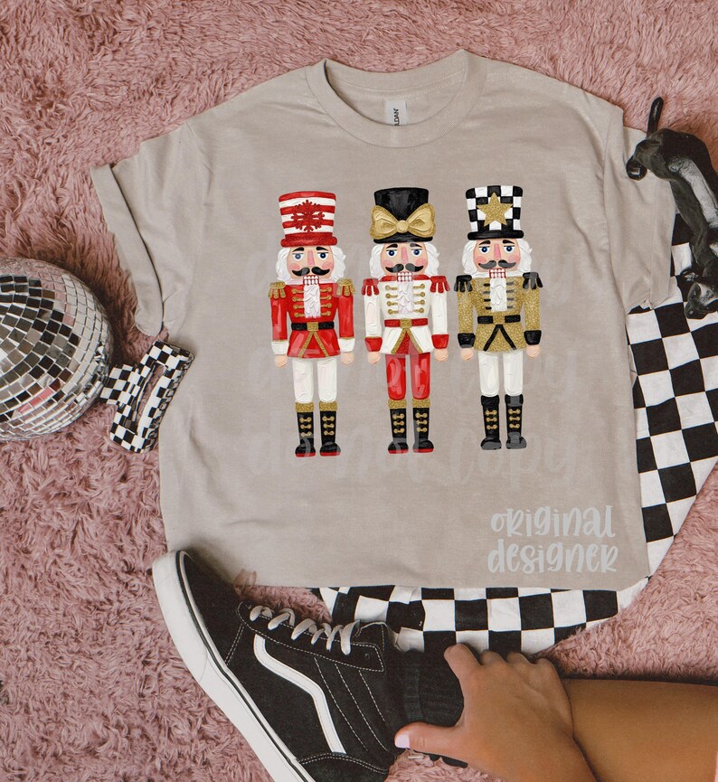 Peut inclure: T-shirt beige avec trois illustrations de casse-noisettes color&eacute;s. Les casse-noisettes portent des uniformes rouges, blancs et dor&eacute;s avec des bottes noires. Le texte "original designer" est imprim&eacute; sur le t-shirt.