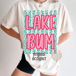 May include: Cream-colored t-shirt with the words "LAKE BUM" in pink, outlined in white, over a teal leopard print design. Below the text is "original designer" in black script. The shirt has short sleeves.