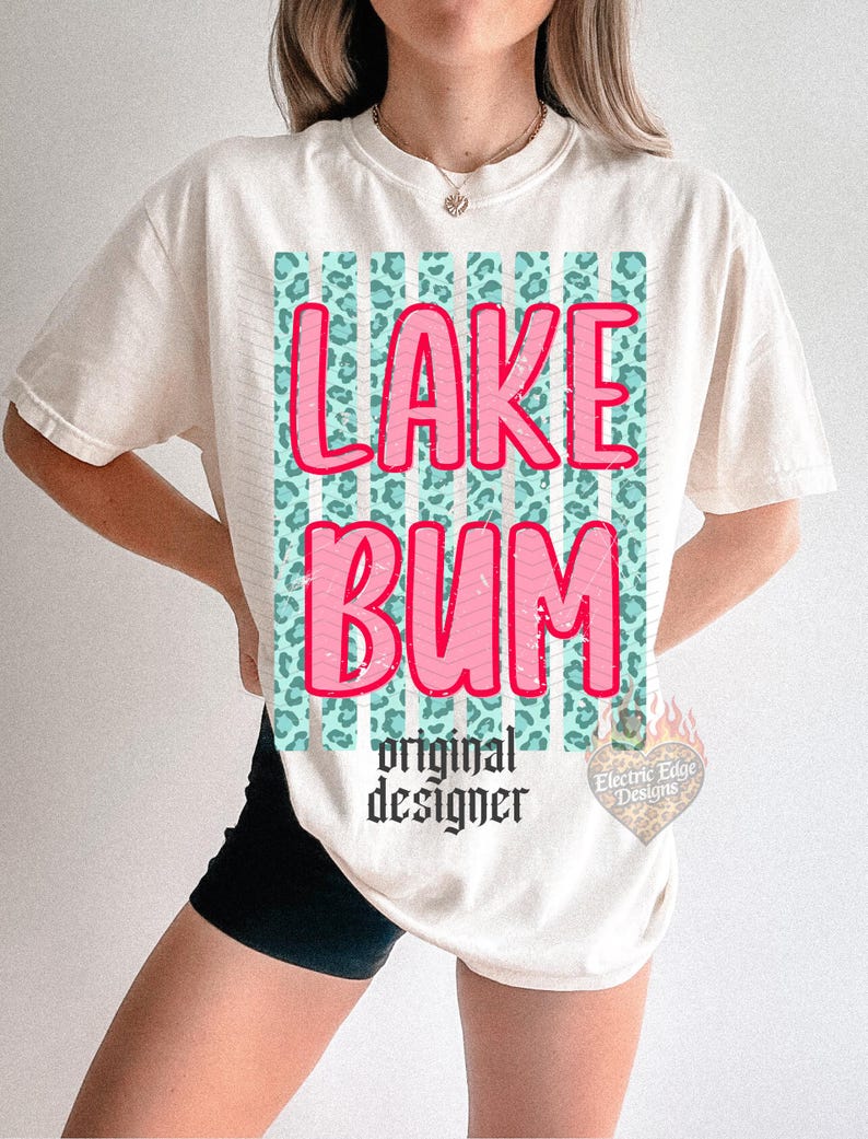 May include: Cream-colored t-shirt with the words "LAKE BUM" in pink, outlined in white, over a teal leopard print design. Below the text is "original designer" in black script. The shirt has short sleeves.