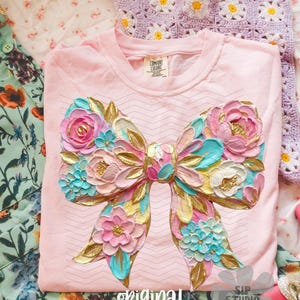 Puede incluir: Una camiseta de manga larga rosa claro con un gran diseño de lazo texturizado. El lazo presenta detalles florales rosas, azules, dorados y blancos. Las palabras "original designer" están impresas en la parte inferior de la camiseta.