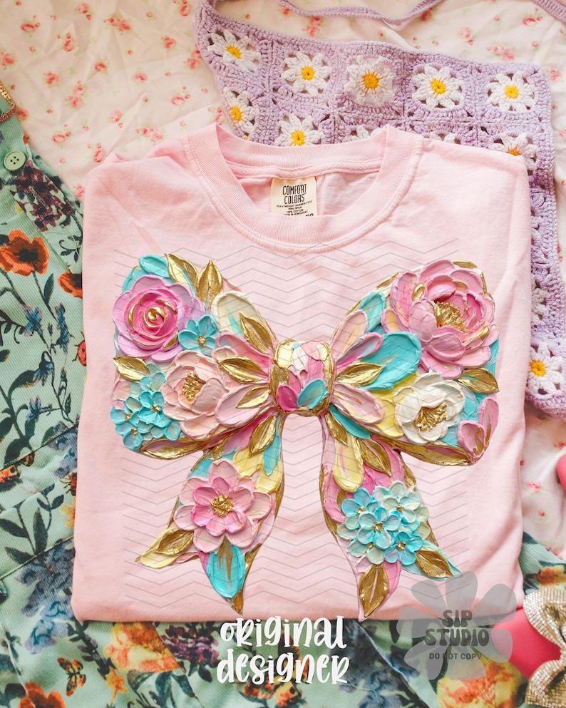 May include: A light pink long-sleeve shirt with a large, textured bow design. The bow features pink, blue, gold, and white floral accents. The words "original designer" are printed at the bottom of the shirt.
