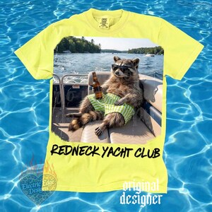 May include: Yellow t-shirt with a graphic of a raccoon wearing sunglasses and green striped shorts, holding a beer bottle on a boat. The text "REDNECK YACHT CLUB" is printed below the graphic.