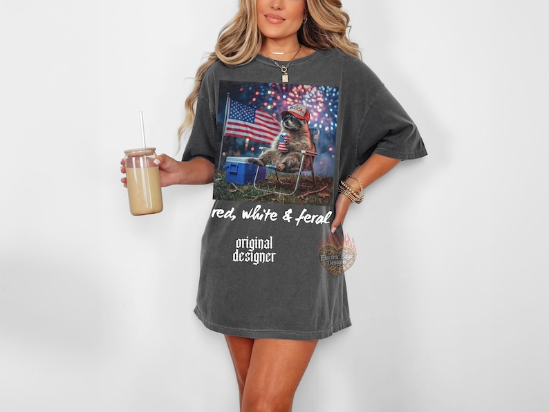 May include: Dark gray t-shirt featuring a graphic of a cat wearing a patriotic hat, sitting in a camping chair with an American flag and fireworks in the background. The shirt has the text "red, white & feral" and "original designer".