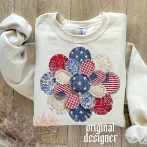 May include: Cream-colored sweatshirt featuring a large, appliqued flower design. The flower is composed of various fabric patches in red, white, and blue patterns, including stars, stripes, and paisley. The text "original designer" is visible.