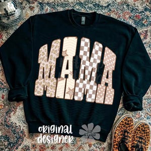 May include: Black sweatshirt with the word "MAMA" in a retro design. The letters feature various patterns, including leopard print, stars, and checkered patterns. The words "original designer" are printed at the bottom.