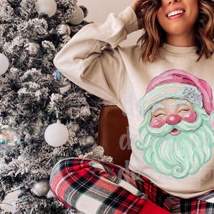 May include: A beige sweatshirt featuring a Santa Claus graphic with a pink hat, mint green beard, and rosy cheeks. The person is wearing red and black plaid pajama pants. A decorated Christmas tree is in the background.