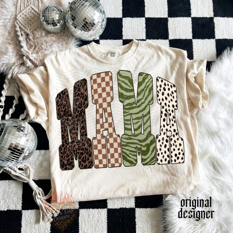 May include: A cream-colored t-shirt featuring the word MAMA in large block letters. Each letter has a distinct pattern: leopard print, brown and white checkerboard, green zebra stripes, and black and white dalmatian spots on a light background.