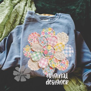 May include: A light blue sweatshirt with a large, colorful flower applique. The flower is made of various patterned fabric circles in shades of pink, yellow, blue, and white. The text "original designer" is printed in white script on the bottom right.