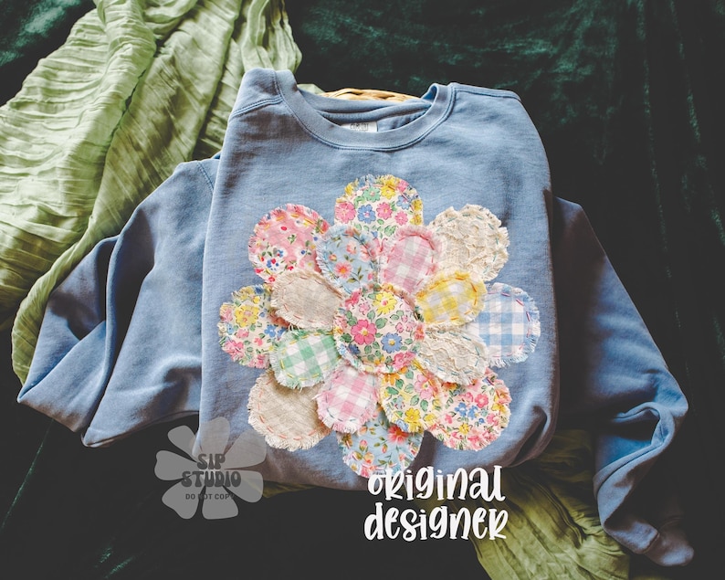 May include: A light blue sweatshirt with a large, colorful flower applique. The flower is made of various patterned fabric circles in shades of pink, yellow, blue, and white. The text "original designer" is printed in white script on the bottom right.
