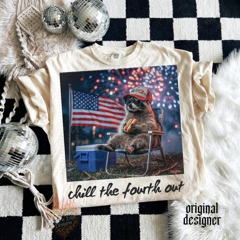 K&ouml;nnte beinhalten: Cremefarbenes T-Shirt mit einem Waschb&auml;ren, der eine Sonnenbrille und einen roten Hut mit der Aufschrift "America" tr&auml;gt und einen Hotdog h&auml;lt. Der Waschb&auml;r sitzt in einem Stuhl vor einer amerikanischen Flagge und Feuerwerk. Der Text "chill the fourth out" steht darunter.