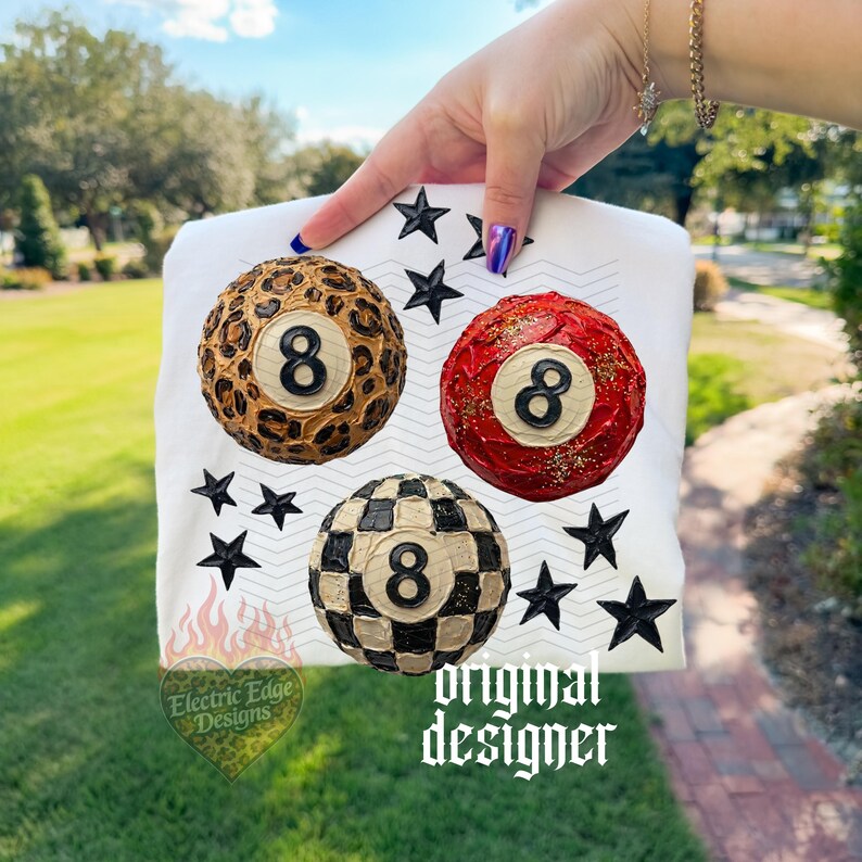 May include: White t-shirt featuring three stylized 8-ball designs: leopard print, red with gold accents, and black and white checkered. Black star accents surround the balls. The text "Electric Edge Designs" and "original designer" are also visible.