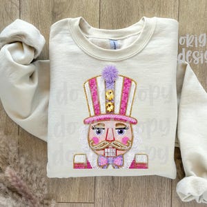 May include: Cream-colored sweatshirt featuring an embroidered nutcracker design. The nutcracker has a pink and white striped hat, a purple pom-pom, and a pink bow tie. The design includes gold accents and a detailed face.