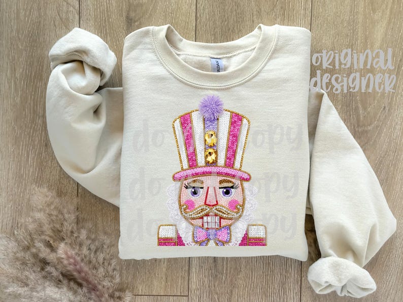 May include: Cream-colored sweatshirt featuring an embroidered nutcracker design. The nutcracker has a pink and white striped hat, a purple pom-pom, and a pink bow tie. The design includes gold accents and a detailed face.