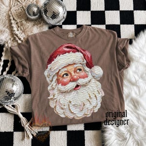 Puede incluir: Una camiseta marr&oacute;n de manga corta con un gr&aacute;fico central de Santa Claus con una espesa barba blanca y un gorro rojo. La ilustraci&oacute;n tiene un estilo de pintura texturizada. El texto original designer est&aacute; impreso en negro abajo a la derecha.