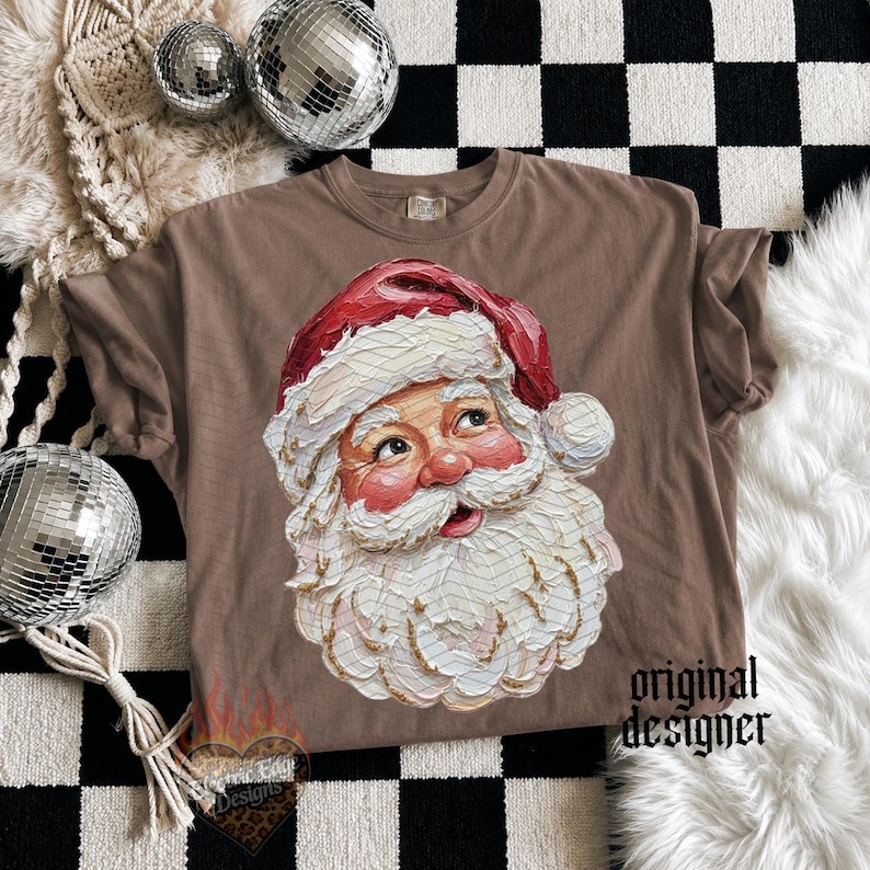 Puede incluir: Una camiseta marr&oacute;n de manga corta con un gr&aacute;fico central de Santa Claus con una espesa barba blanca y un gorro rojo. La ilustraci&oacute;n tiene un estilo de pintura texturizada. El texto original designer est&aacute; impreso en negro abajo a la derecha.