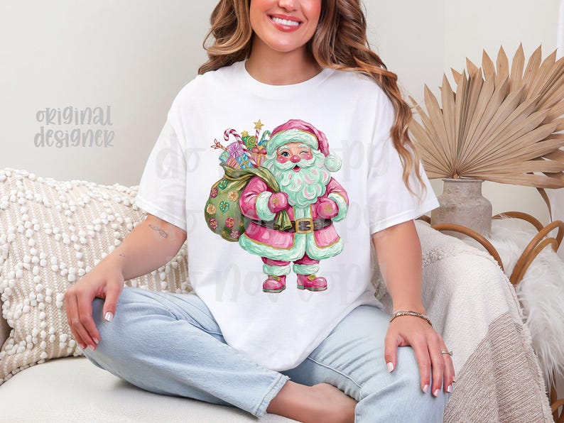 May include: White t-shirt with a watercolour illustration of Santa Claus in pink and green, carrying a sack of gifts. The design includes candy canes and wrapped presents, ideal for the festive season.
