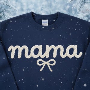 May include: Navy blue sweatshirt with the word "mama" in a cream-colored, textured font. A cream-colored bow is below the word. The sweatshirt has a crew neck and a tag that says "Gildan". Snowflakes are in the background.