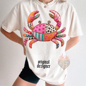 May include: Cream-colored t-shirt featuring a colorful crab design. The crab is made up of various patterned patches in pink, orange, green, and leopard print. The text "original designer" is printed on the shirt.