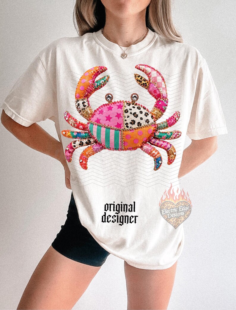 May include: Cream-colored t-shirt featuring a colorful crab design. The crab is made up of various patterned patches in pink, orange, green, and leopard print. The text "original designer" is printed on the shirt.