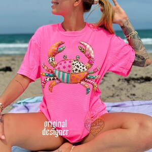 May include: A bright pink t-shirt featuring a colorful crab design. The crab is made of various patterned fabrics, including stars, polka dots, and stripes. The shirt has a relaxed fit and a subtle chevron pattern. The words "original designer" are printed on the shirt.