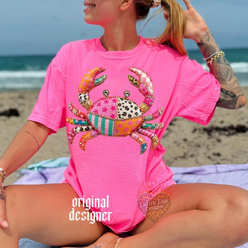 May include: A bright pink t-shirt featuring a colorful crab design. The crab is made of various patterned fabrics, including stars, polka dots, and stripes. The shirt has a relaxed fit and a subtle chevron pattern. The words "original designer" are printed on the shirt.