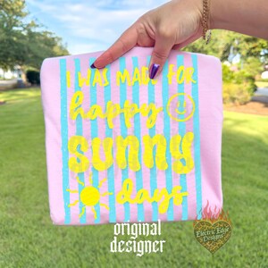 May include: Pink t-shirt with blue vertical stripes and yellow text that reads "I WAS MADE FOR happy SUNNY days" with a sun and smiley face graphic. The words "original designer" are at the bottom.