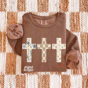 May include: A brown long-sleeve sweatshirt featuring three cross graphics across the chest. The first cross has a blue floral pattern, the second a beige checkered pattern, and the third a pattern of small blue crosses. Text reads original designer.