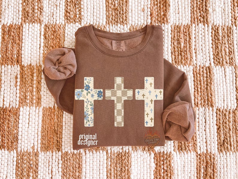May include: A brown long-sleeve sweatshirt featuring three cross graphics across the chest. The first cross has a blue floral pattern, the second a beige checkered pattern, and the third a pattern of small blue crosses. Text reads original designer.
