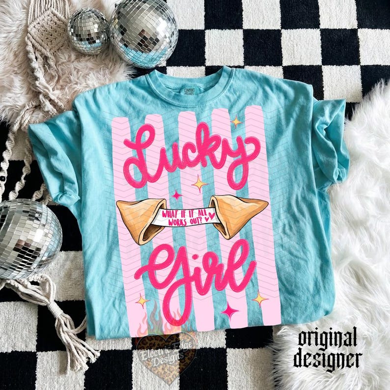 May include: A turquoise t-shirt featuring a pink design with the words "Lucky Girl." The design includes a fortune cookie with the text "What if it all works out?" and pink vertical stripes. The shirt has short sleeves.