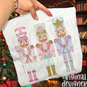 May include: White pillowcase featuring three colorful nutcracker illustrations. Each nutcracker is dressed in pastel pink, mint green, and lavender, with gold accents. One holds a lollipop, and all have detailed uniforms.