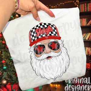 May include: White pillow featuring a graphic of Santa Claus wearing sunglasses with star-shaped lenses. Santa has a white beard and is wearing a red and black checkered hat with the word "Sleigh" on it. The pillow is held by a person.